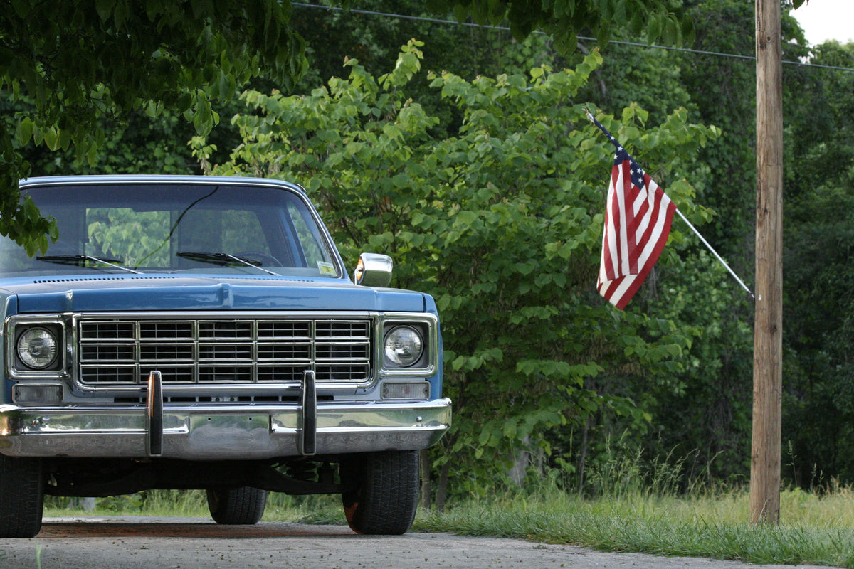 A blue vintage pickup truck parked outdoors on a road with green trees and bushes behind it, and an American flag hanging from a utility pole on the right side of the frame.